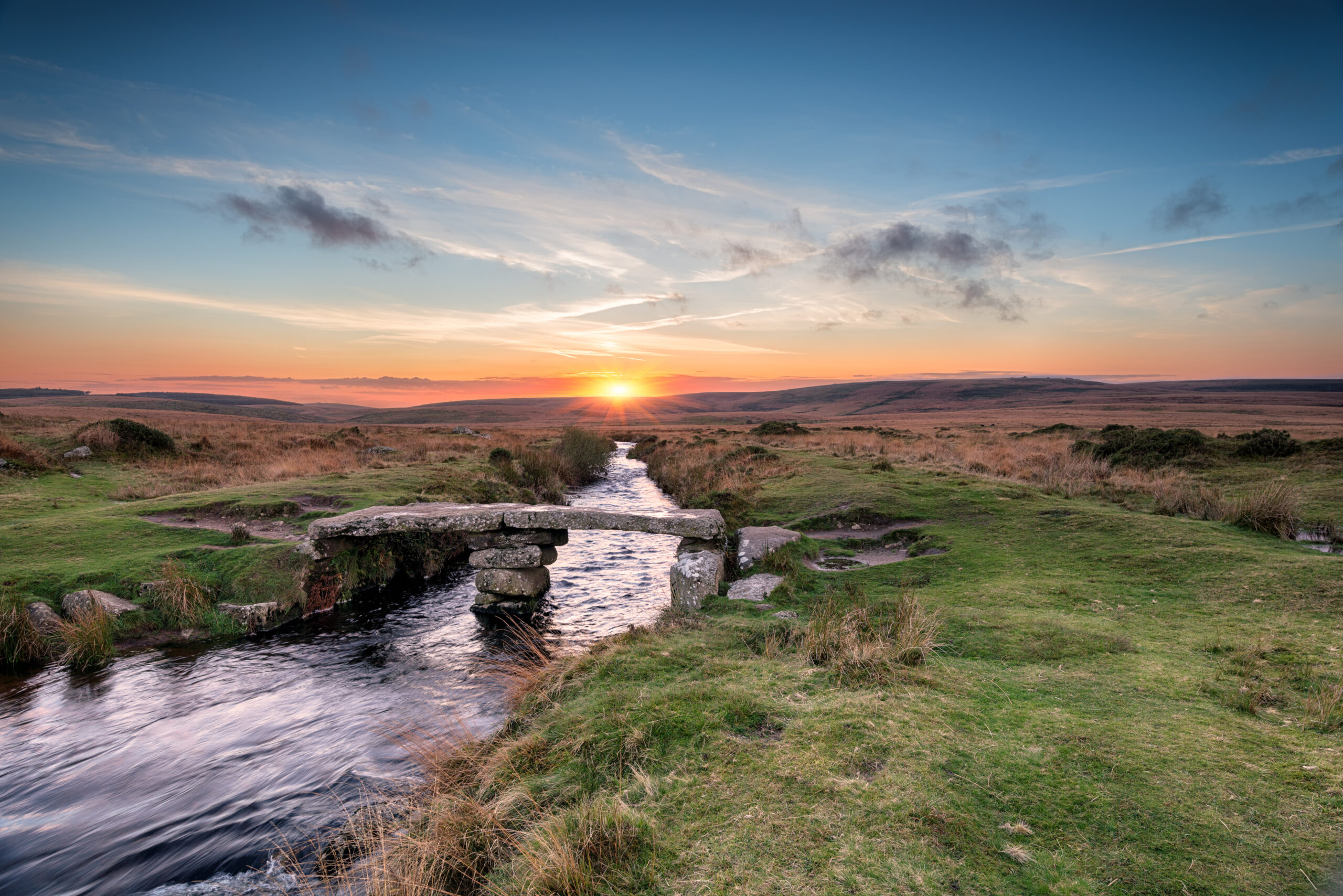 A landscape shot over Dartmoor with a river running through the image and a bridge going over the river. The sun is setting in the background.